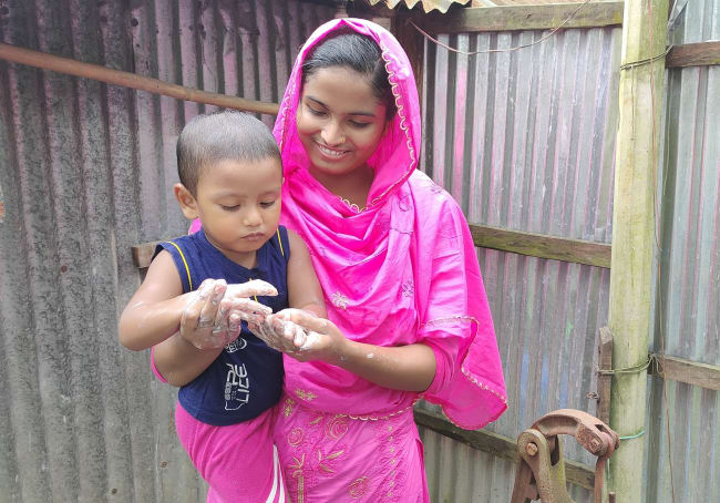 Health leader Sabikunnahar, wearing a bright pink traditional outfit, smiles while helping a young child wash their hands with soap and water outdoors beside a corrugated tin wall.