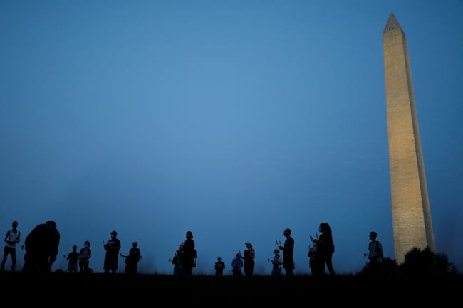 They are informally honored—but are they officially counted? Activists stand at sunrise to memorialize victims of the coronavirus, near the Washington Monument in Washington, DC on August 27, 2020. The photo shows the monument in the thin morning lite with a silhouette line of people standing in the foreground. REUTERS/Tom Brenner