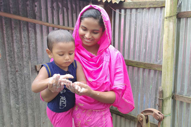 Health leader Sabikunnahar, wearing a bright pink traditional outfit, smiles while helping a young child wash their hands with soap and water outdoors beside a corrugated tin wall.