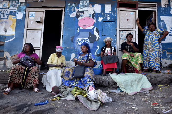 Traders sit near a deserted crossing point between Goma, Democratic Republic of Congo and Rwanda amid concerns about the spread of coronavirus on March 23, 2020. The photo shows a number of women sitting in front of a storefront painted blue and adorned by a large cartoon figure of a cow giving a thumb's up. REUTERS/Olivia Acland 