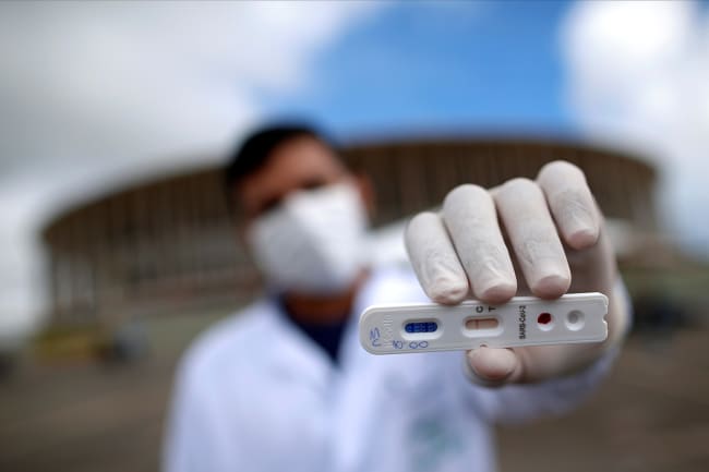 A medical professional shows a negative test for novel coronavirus in front of Mane Garrincha Stadium, amid the coronavirus disease (COVID-19) outbreak, in Brasilia, Brazil, on April 21, 2020. The photo shows a health worker holding a test at the camera with a stadium in the background. the photographer is using a telephoto/macro lens so that everything is blurred out in the shot, including the man, with the exception of the small plastic cassette-like test.  REUTERS/Ueslei Marcelino