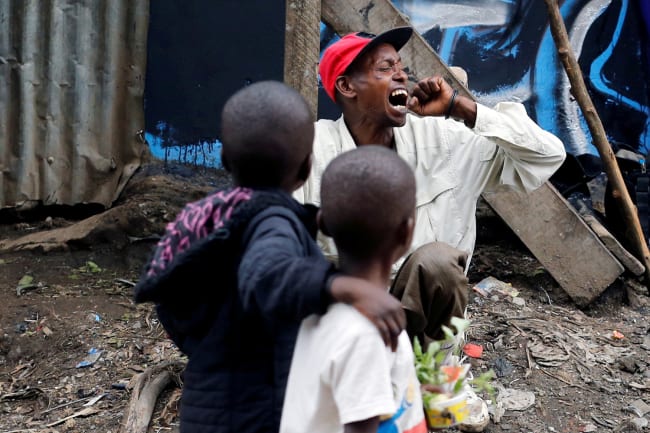 A man coughs as children walk past roadside, during the coronavirus pandemic in Nairobi, Kenya April 19, 2020. This is a powerful image of two children looking at a man who is coughing into his had with his head turned. REUTERS/Thomas Mukoya