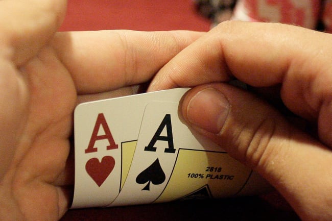 Photo shows a pair of hands holding up the corners of two cards to reveal a pair of aces. A large stack of chips is in the background. 