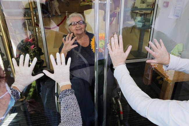 A resident named Mrs. Oury reacts as her daughters wave behind a glass during a visit at "Les Figuiers" retirement home amid the coronavirus outbreak in Villeneuve-Loubet, France, on April 23, 2020. The photo shows a woman from behind glass while two others stand with their backs to the camera looking in. They all have their hands up. REUTERS/Eric Gaillard