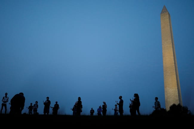 They are informally honored—but are they officially counted? Activists stand at sunrise to memorialize victims of the coronavirus, near the Washington Monument in Washington, DC on August 27, 2020. The photo shows the monument in the thin morning lite with a silhouette line of people standing in the foreground. REUTERS/Tom Brenner