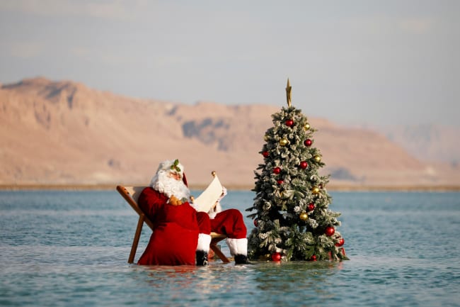 A man, wearing a red Santa Claus costume complete with a hat, beard, and bag full of toys, holds a scroll as he poses for a picture while sitting next to a decorated Christmas tree on a salt formation in the Dead Sea, near Ein Bokeq, Israel on November 15, 2020.