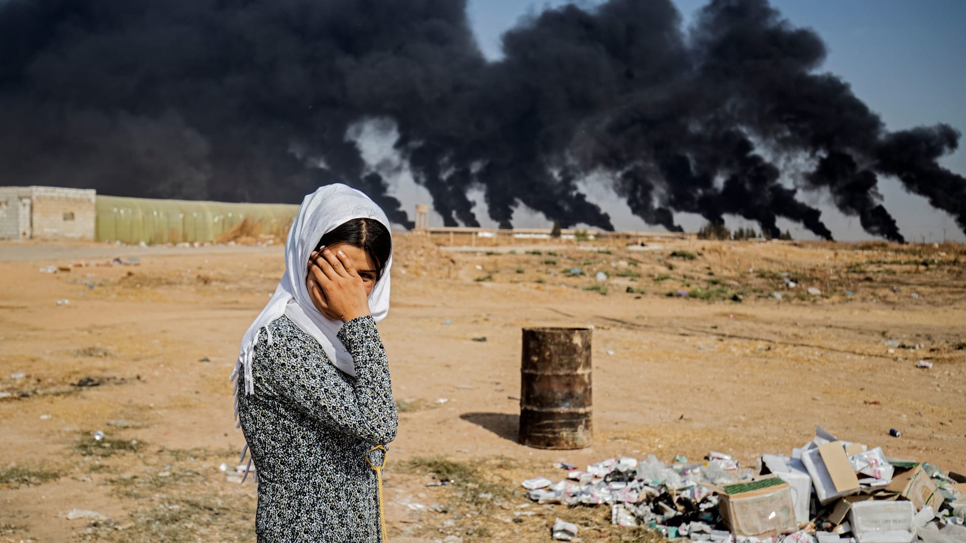 <p>A woman covers her face as she stands on the outskirts of a town near the Syria-Turkey border, while smoke plumes meant to decrease visibility for Turkish warplanes billow from tire fires during a cross-border offensive, on October 16, 2019.</p>
