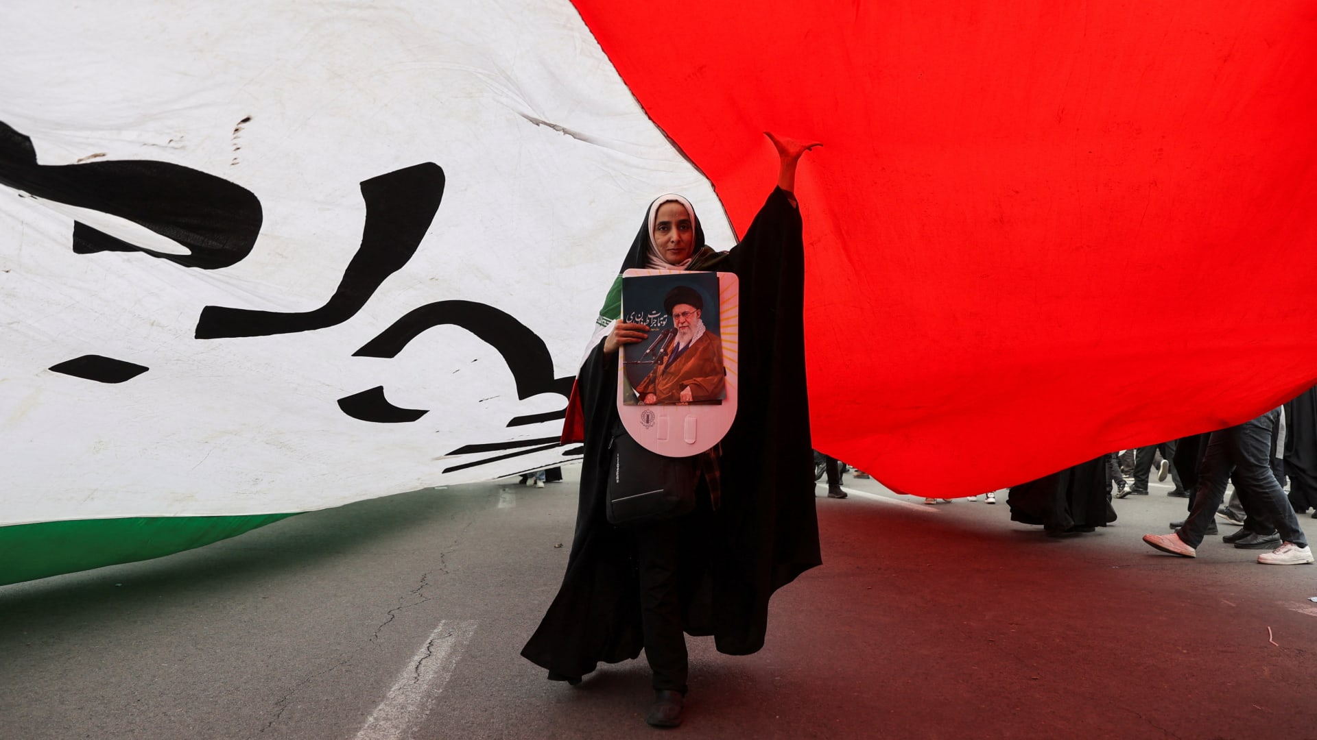 An Iranian woman holding a poster depicting Iran's Supreme Leader Ayatollah Ali Khamenei walks under a large flag during the forty-seventh anniversary of the Islamic Revolution in Tehran, Iran on February 11, 2026. Majid Asgaripour/West Asia News Agency via Reuters