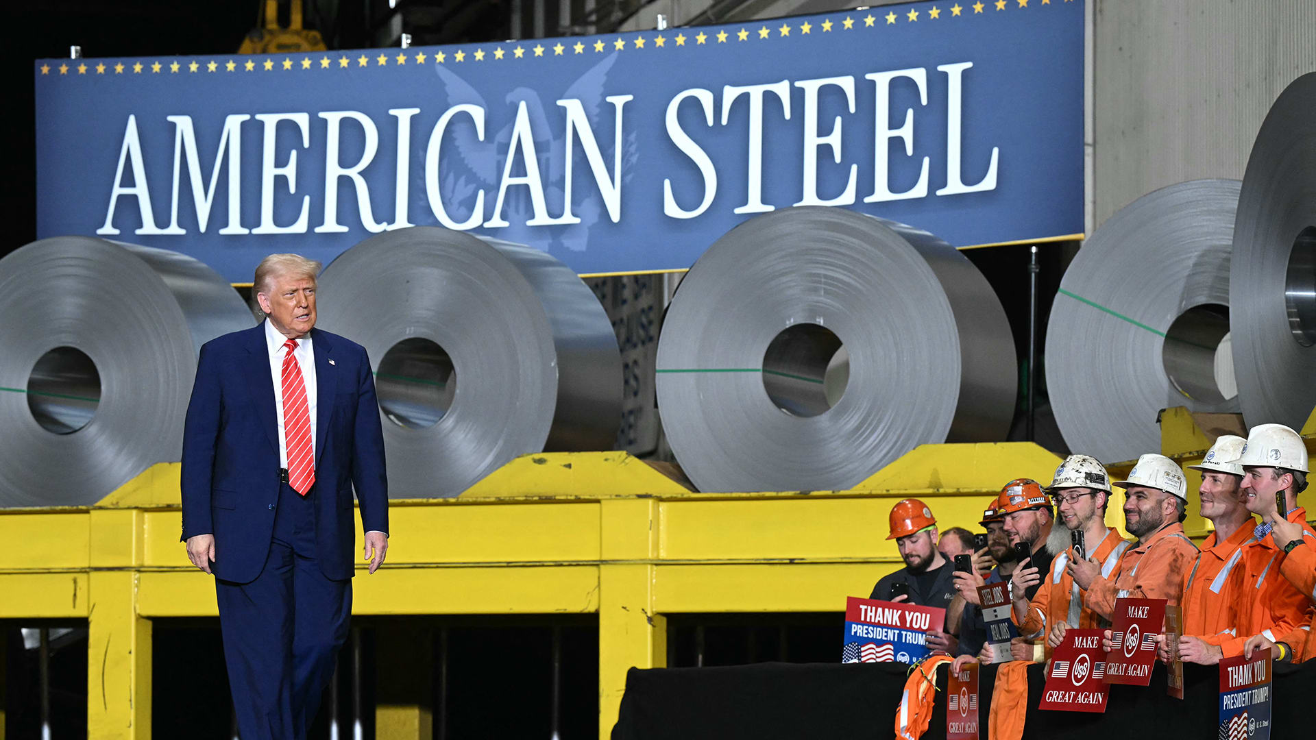 Photo of president trump standing in front of a banner and a pile of steel rolls to mark deal between Nippon Steel and US Steel