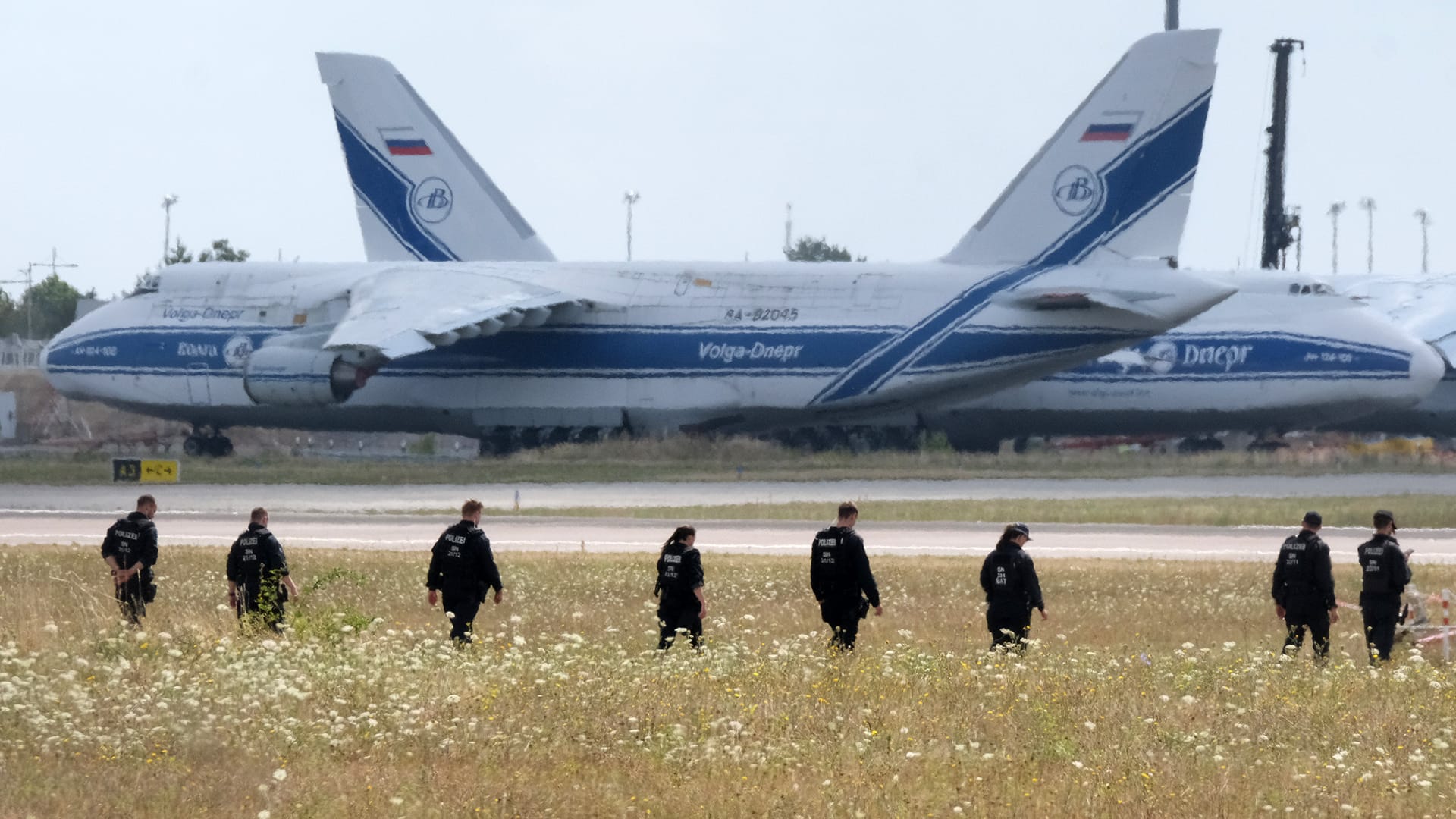 A line of police officers walks through a field of tall grass toward a runway at Leipzig Airport.
