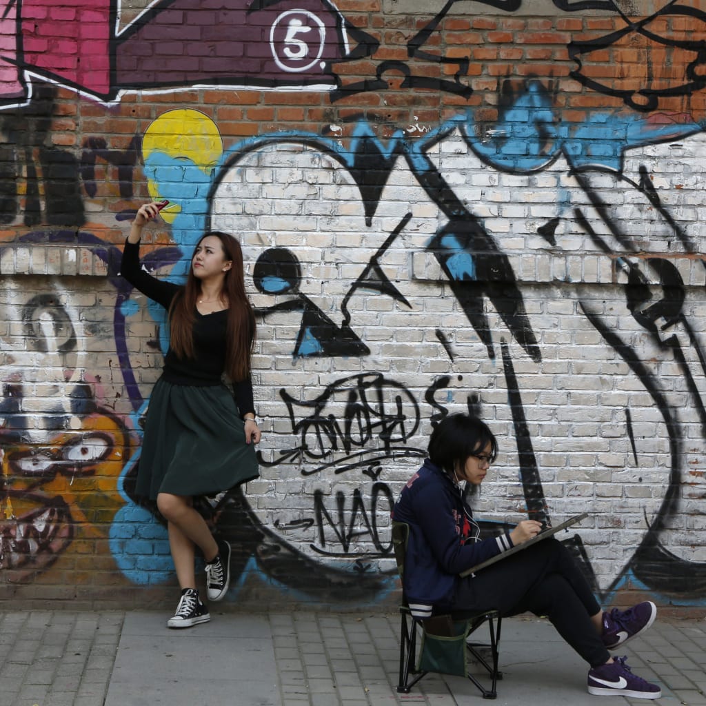 <p>A woman poses for her self-portrait with her mobile phone while another works on a painting in front of a graffiti during the 2013 Beijing 798 Art Festival at the 798 Art Zone in Beijing October 8, 2013. The art zone, originally an unused factory, was tra</p>
