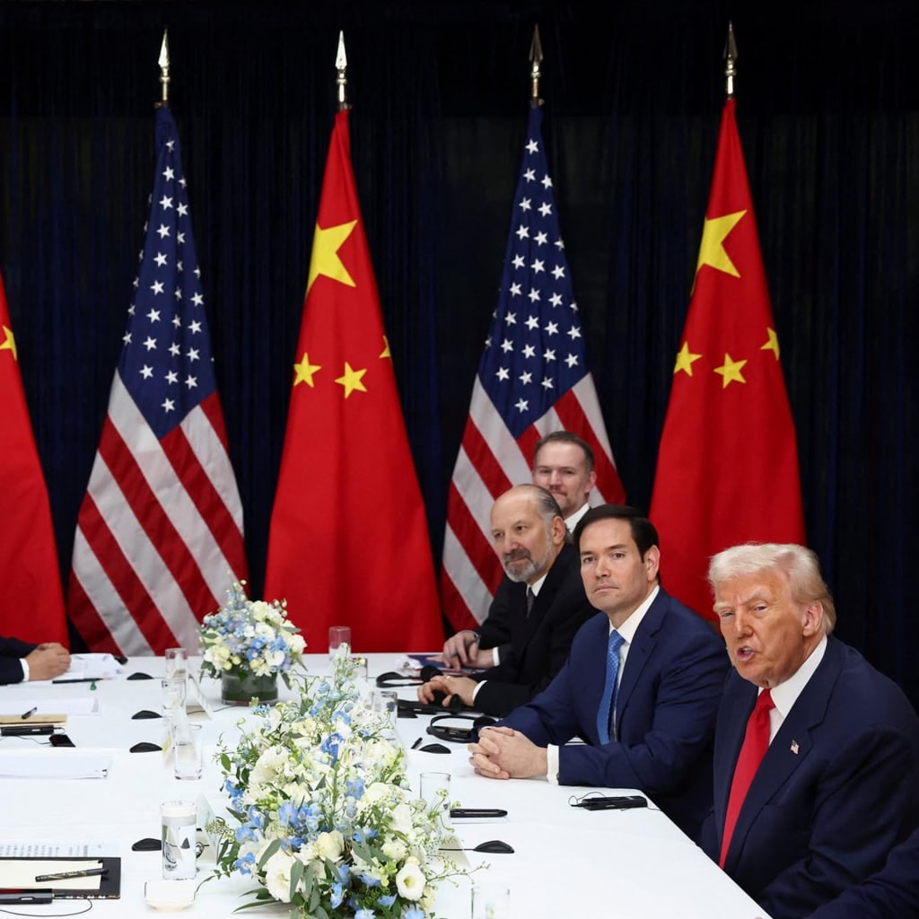 President Trump and President Xi sit across from the table from each other. Other men sit beside them. Chinese and American flags stand in the background