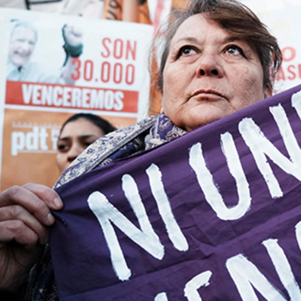 A demonstrator holds a purple piece of fabric that reads "Ni Una Menos", translating to "Not one less", at a protest as others hold various signs up around her.