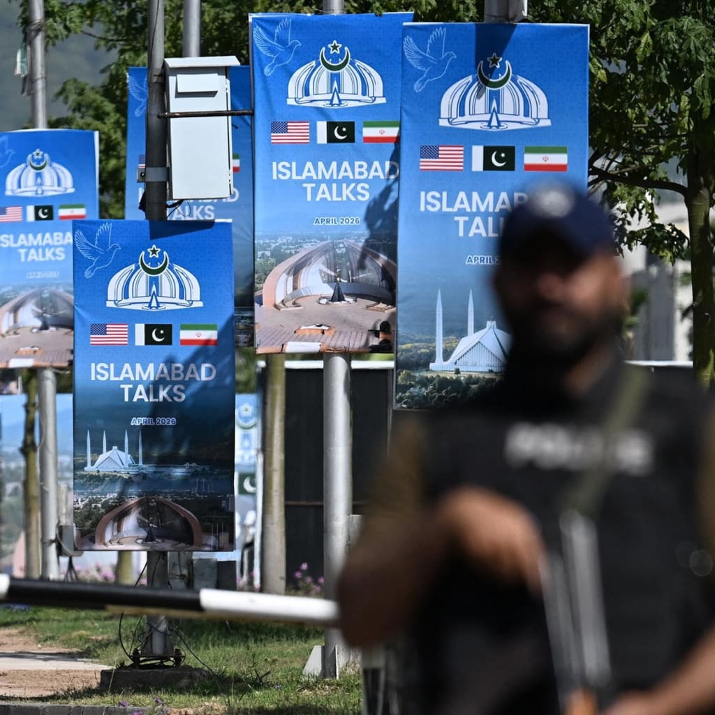 Two security guards stand armed in front of a road that has several banners displaying signage that read "Islamabad talks April 2026".