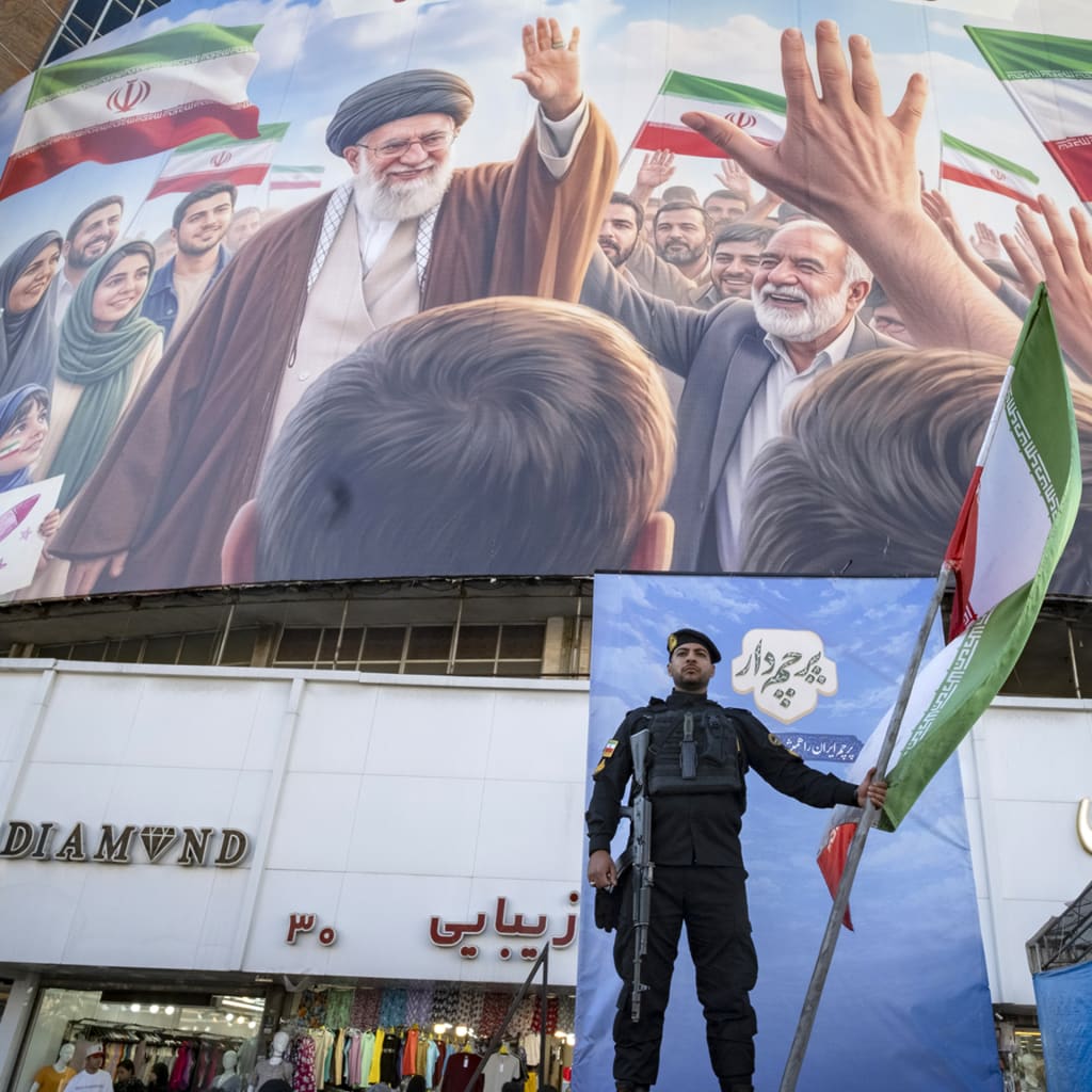 An Iranian police officer holds a national flag on a stage alongside a Persian script reading 'Flag bearer', under a giant billboard depicting an AI-generated portrait of Iran's late Supreme Leader Ayatollah Ali Khamenei in downtown Tehran, Iran, on April 23, 2026.