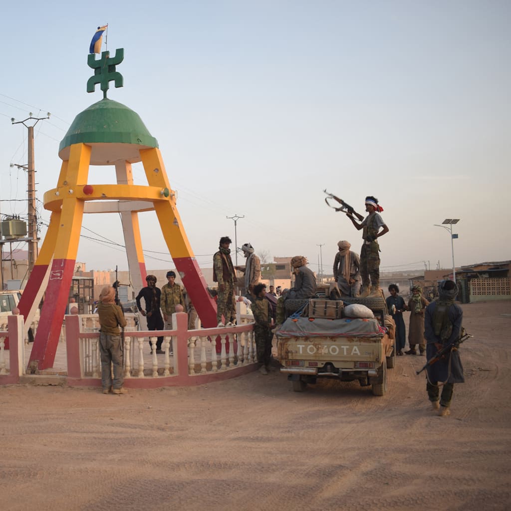 Soldiers gather on the dirt in front of a red, yellow, and green monument, with one soldier standing with his gun pointed up while on top of a truck.