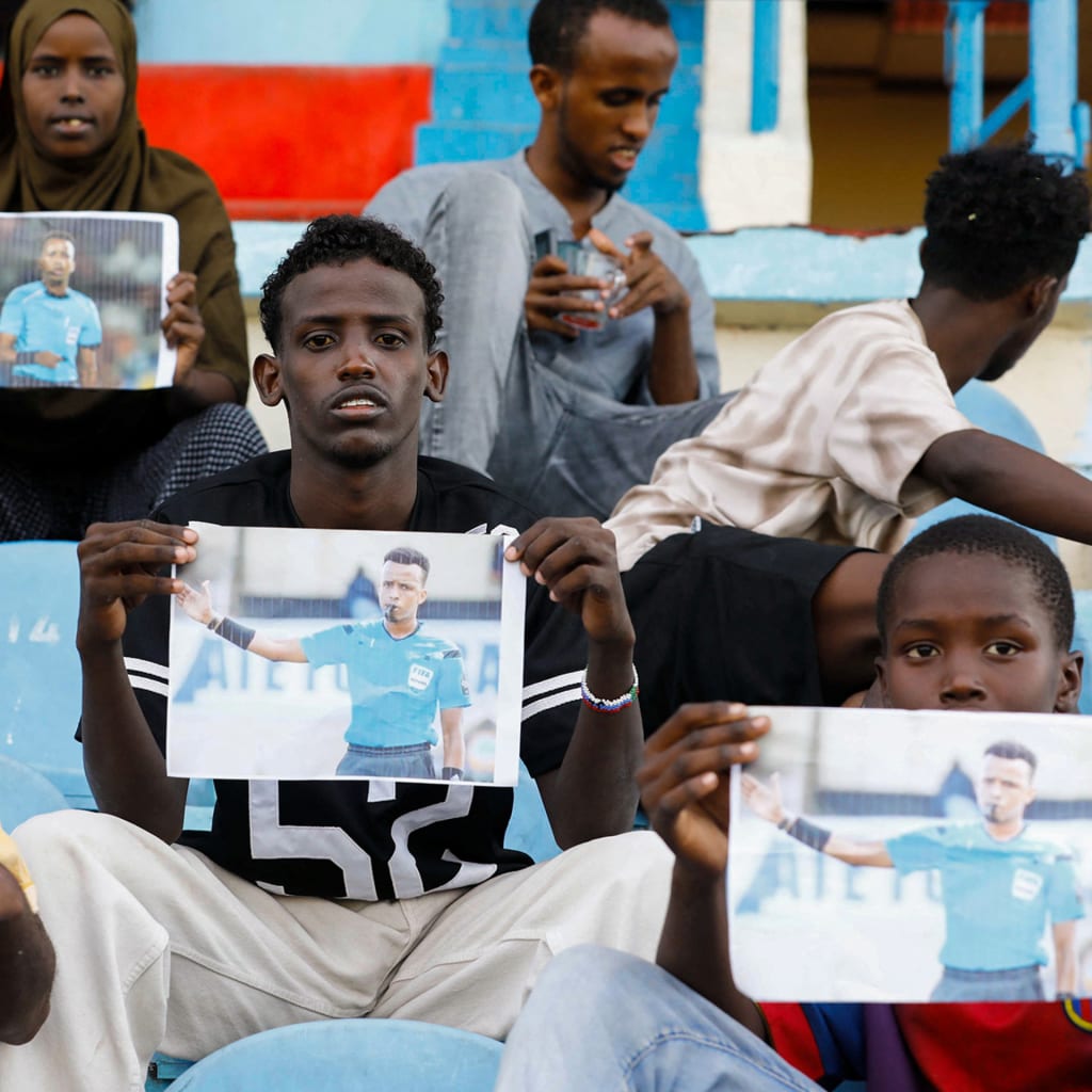 Somali soccer fans sit in a stadium, holding photos of referee Omar Abdulkadir Artan, who had been expected to officiate matches at the 2026 FIFA World Cup but was refused entry to the United States.