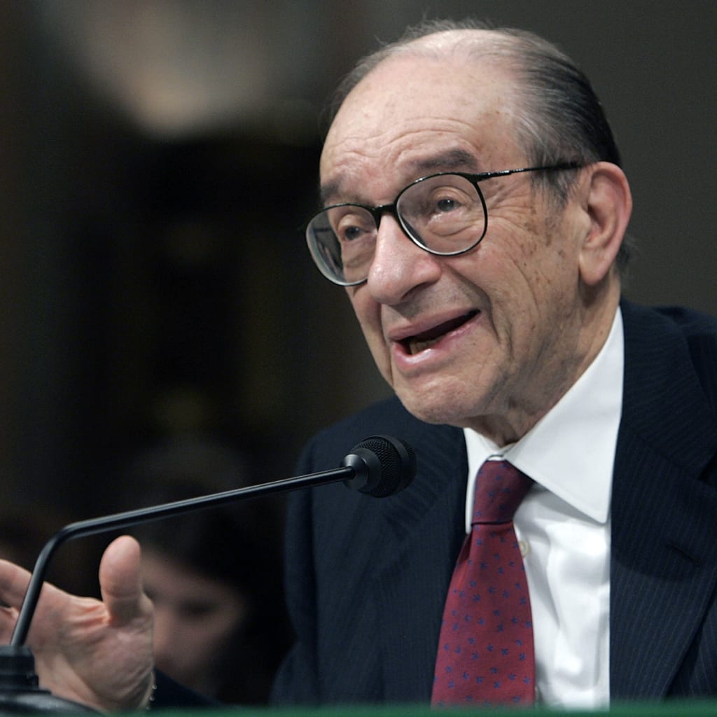 Alan Greenspan, dressed in a suit and red tie, gestures while speaking before a microphone.