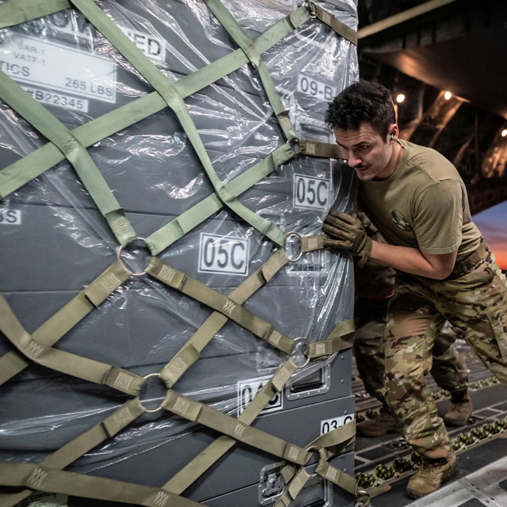 An airman pushes a cargo pallet onto a military transport aircraft. The sunset can be seen in the background.