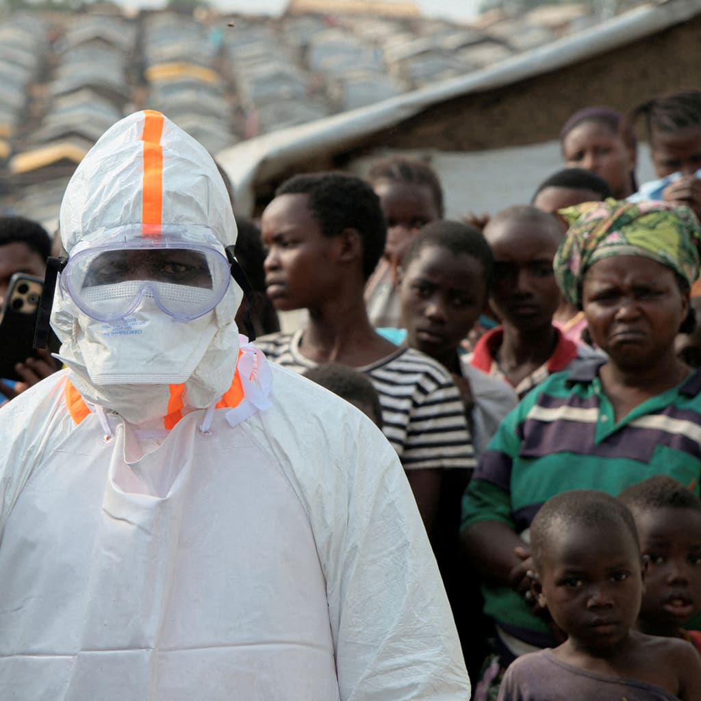 A health worker in a white hazmat suit stands in front of a group of people looking on in a displacement camp.