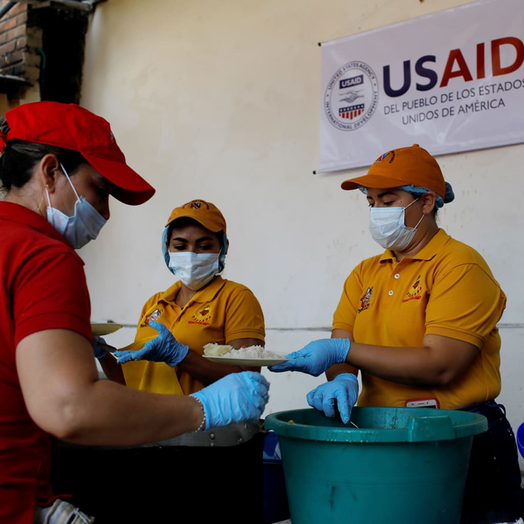 Volunteers serve lunch in a community kitchen set up by USAID and World Food Programme in Cucuta, Colombia. There is a sign behind them that reads, "USAID Del Pueblo de Los Estados Unidos de America".