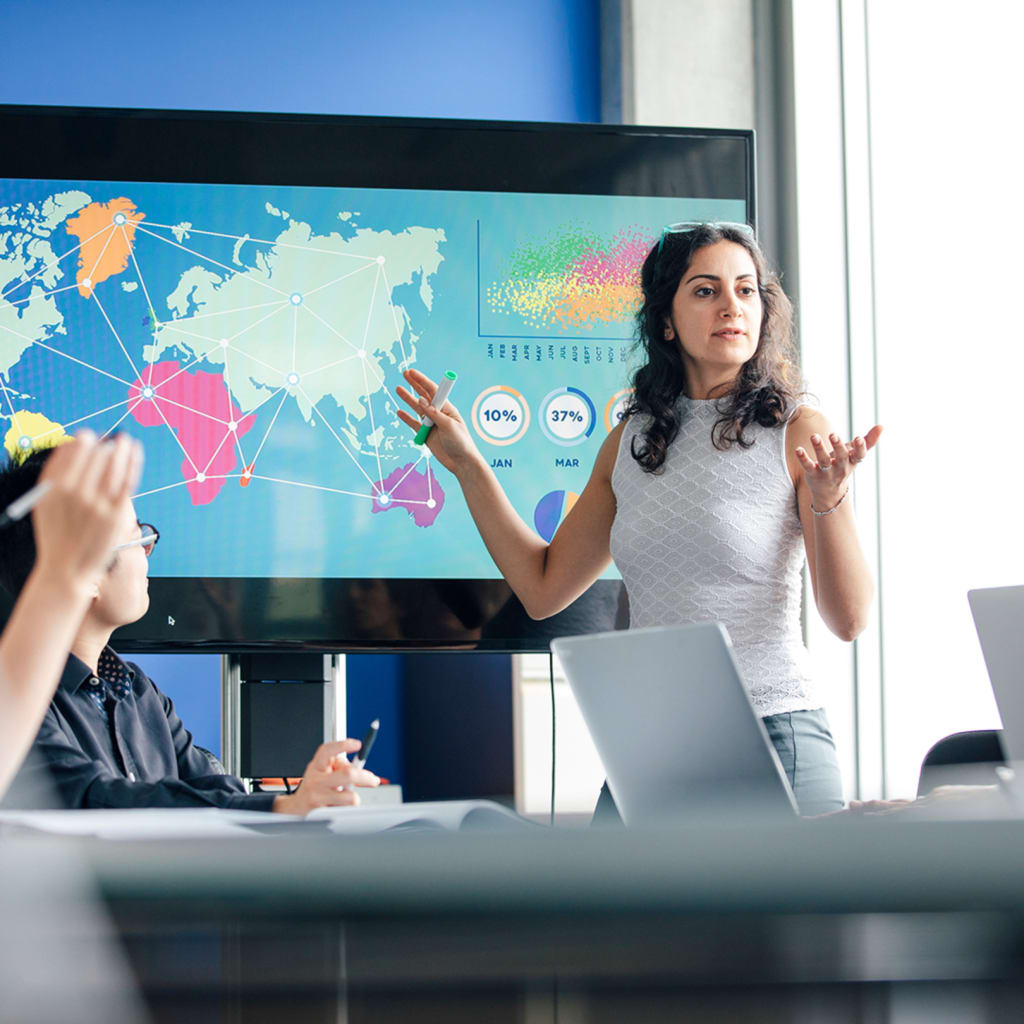 A woman stands in front of a screen that shows a world map. Other are seated at a table listening to her.
