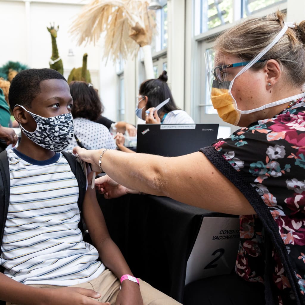 A boy sits in a chair while getting a vaccine from a health worker, both of them wear face masks.