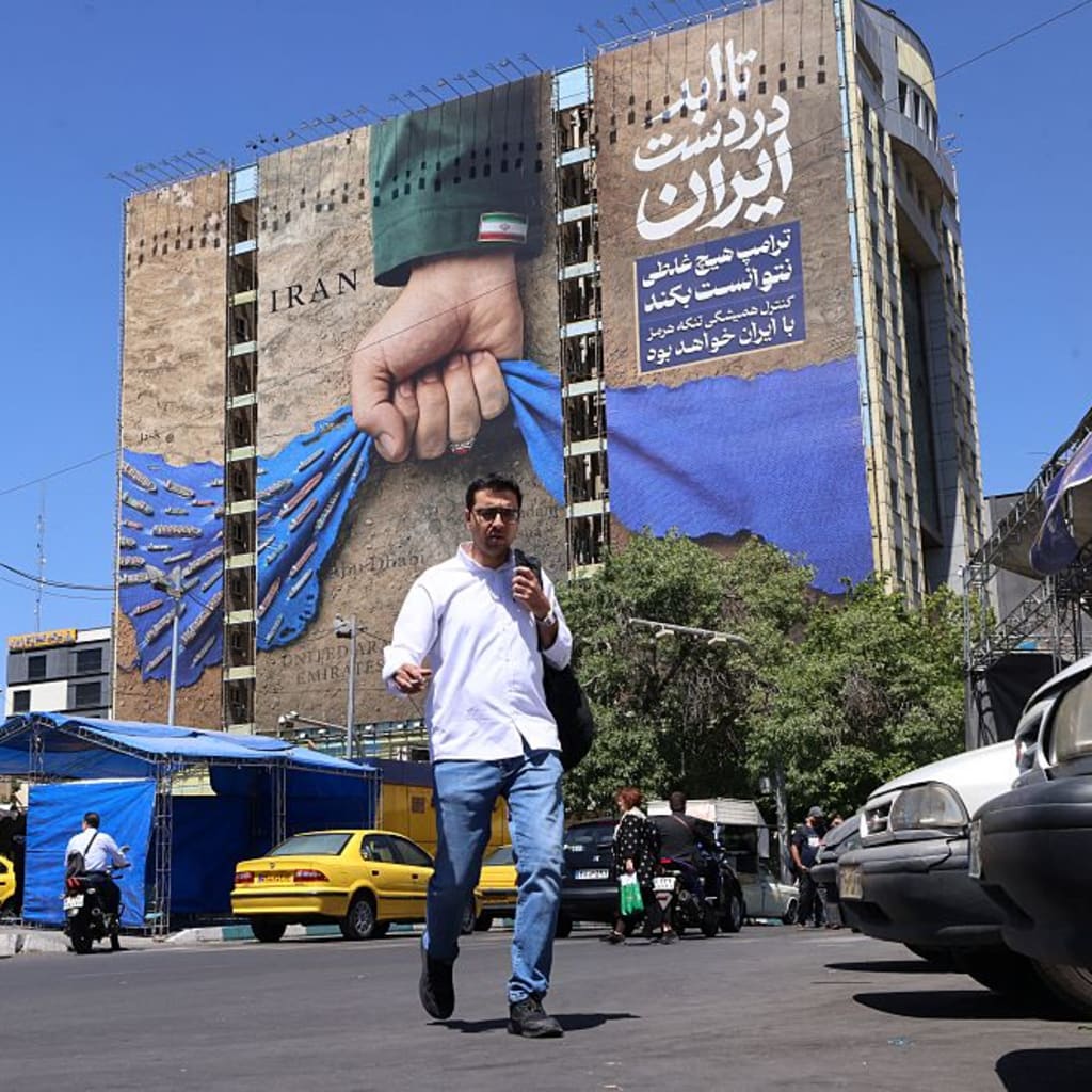 People cross a street past a billboard on the facade of a building depicting the Strait of Hormuz with a caption in Persian reading "Forever in Iran's Hand", at Vanak Square in Tehran on May 25, 2026. I
