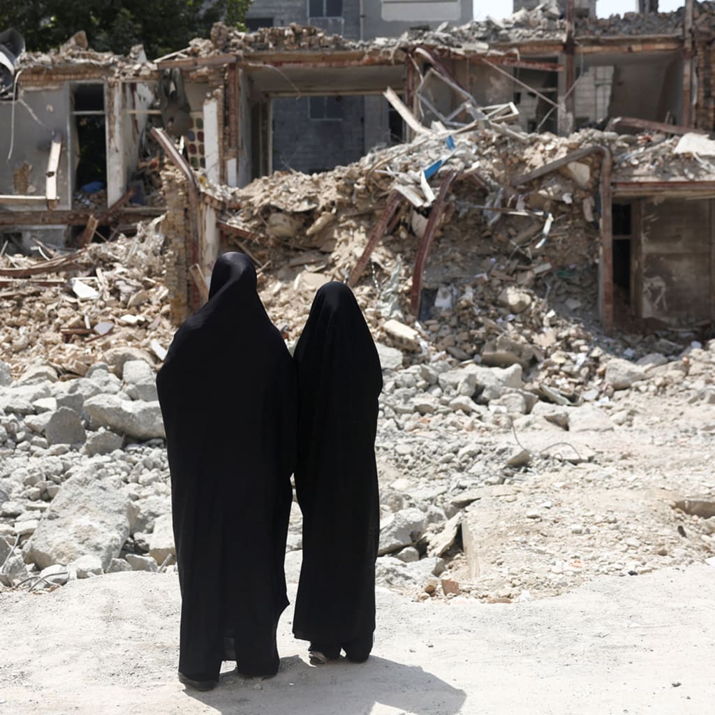 Two Iranian women in burqas face away and look at the rubble from a damaged building.