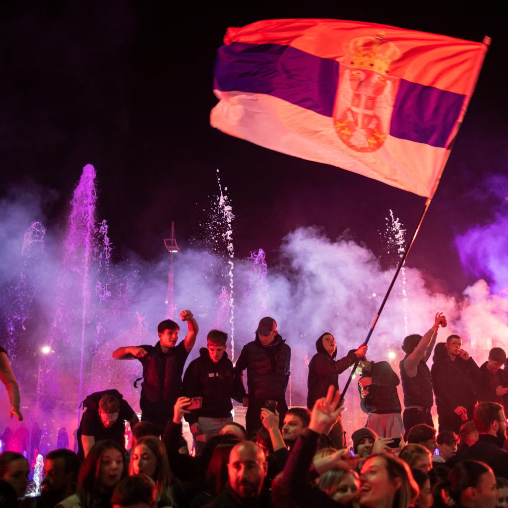 A crowd of people celebrates at night around an illuminated fountain in Belgrade’s Slavija Square, one of them waving a large Serbian flag amid colored smoke.