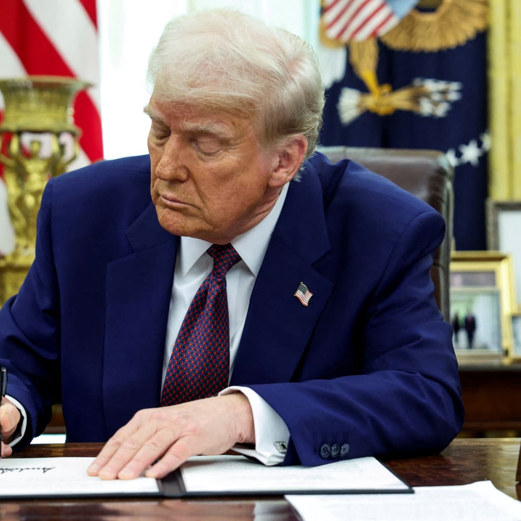 President Donald Trump signs an executive order at the Resolute Desk in the Oval Office, with an American flag and a map behind him.