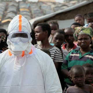 A health worker in a white hazmat suit stands in front of a group of people looking on in a displacement camp.