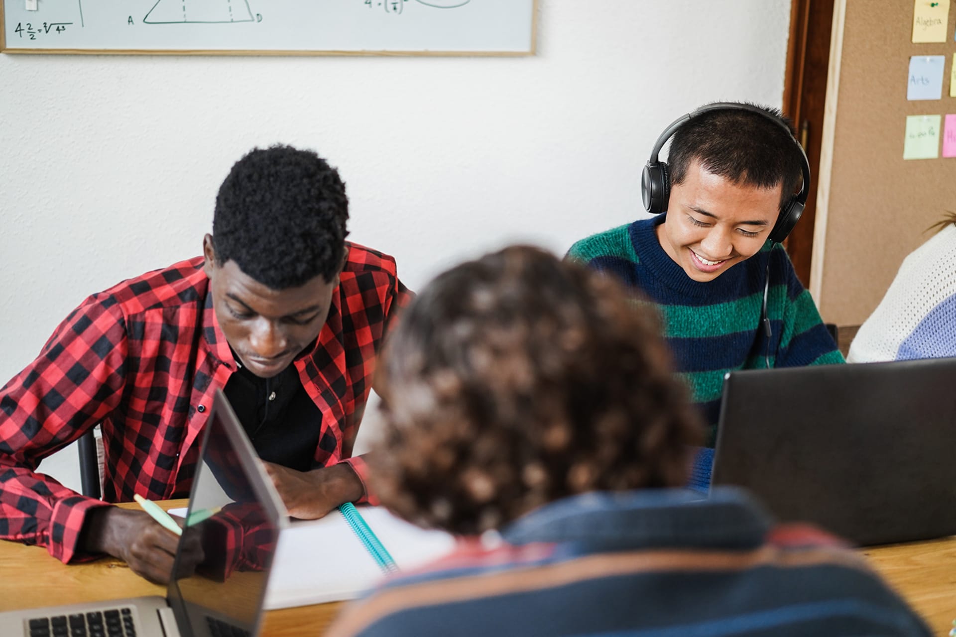 Two students sitting side by side doing work in a classroom. One student has headphones on and is working on a laptop, the other is writing in a notebook.