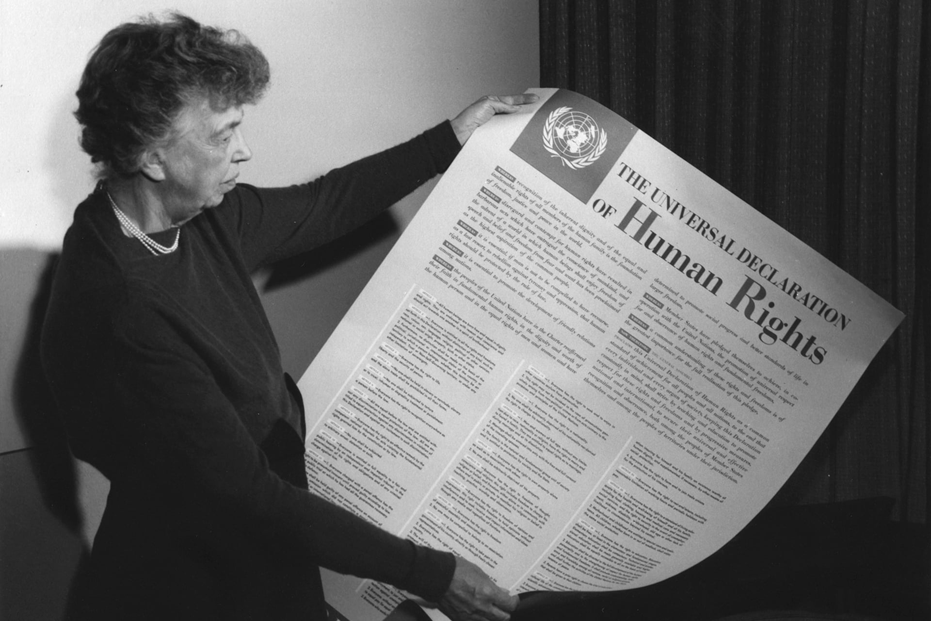 A black and white photo of Eleanor Roosevelt holding a large poster of the Universal Declaration of Human Rights (in English), at Lake Success, New York on November 1949.