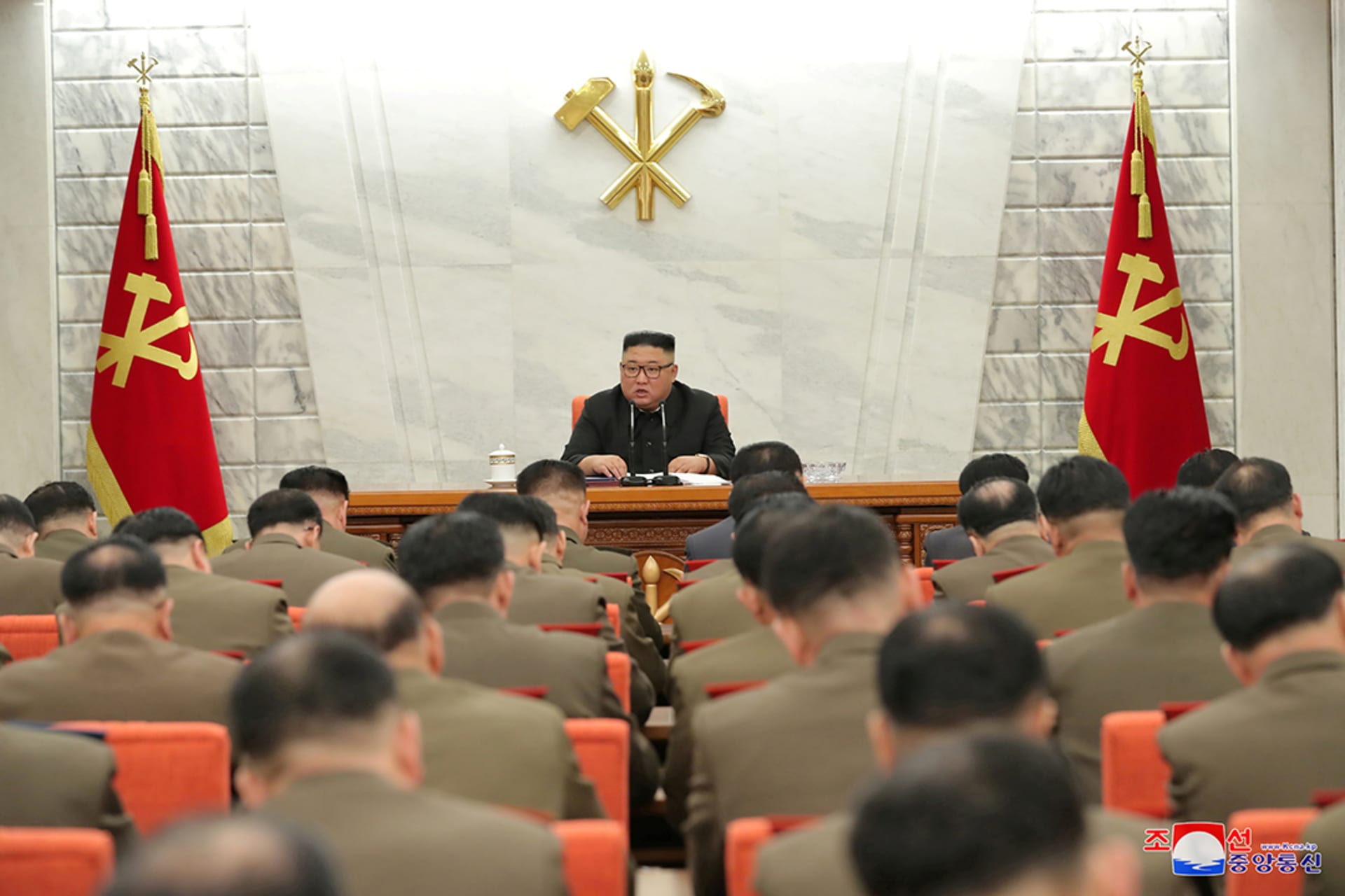 Kim Jong-un wears a white shirt and sits in front of a white wall with red and yelow flags while men sit facing him wearing tan jackets.