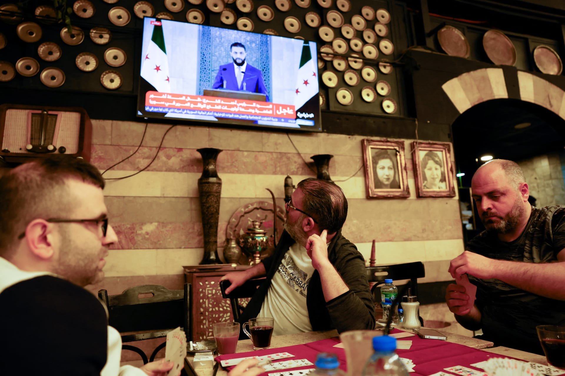 <p>Men play cards at a coffee shop as Syrian President Ahmed al-Sharaa addresses the nation in a televised speech, in Damascus, Syria on May 14.</p>
