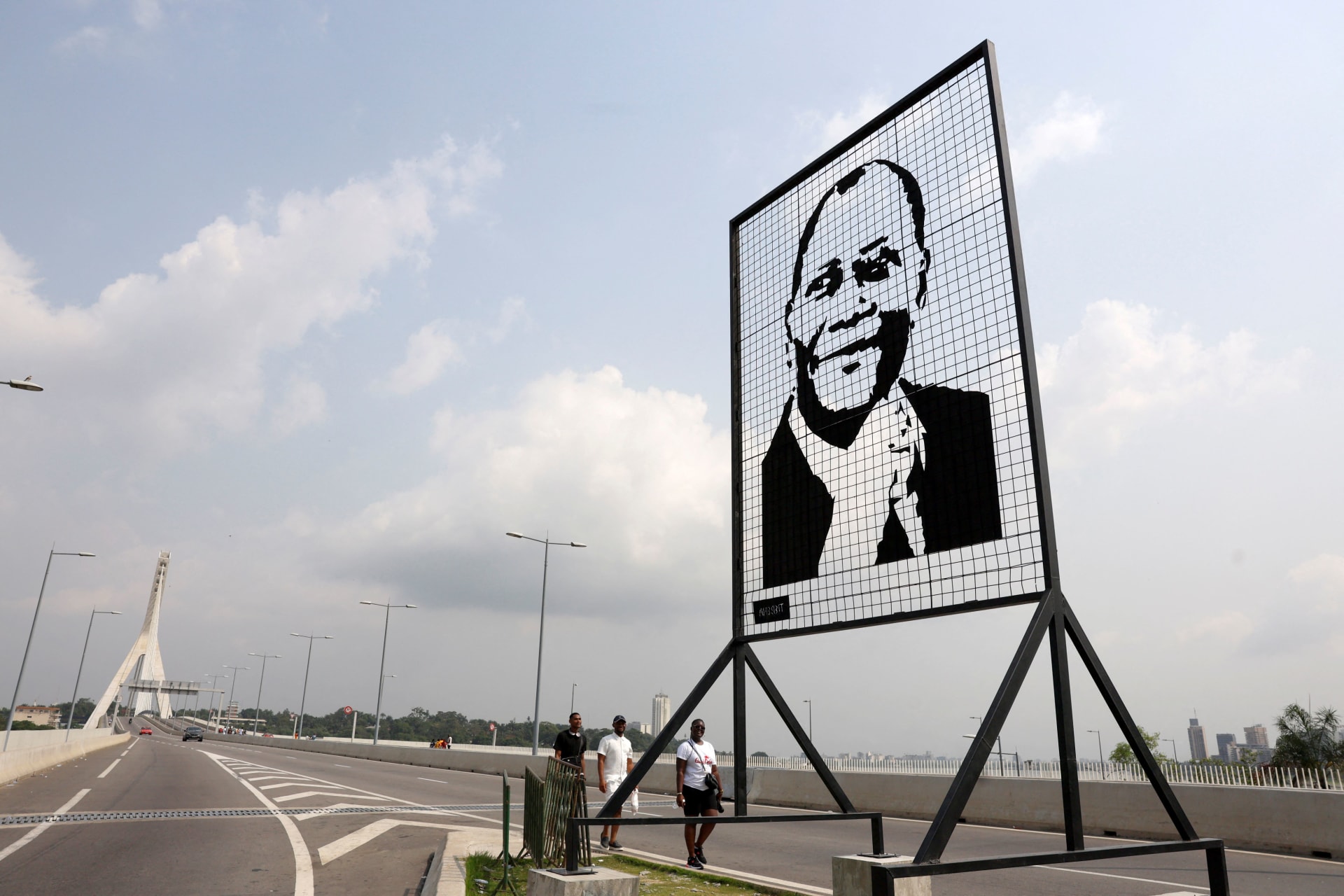 <p>People walk past a portrait of Côte d’Ivoire President Alassane Ouattara at the Alassane Ouattara Bridge in Abidjan, Côte d’Ivoire on January 1, 2025.</p>
