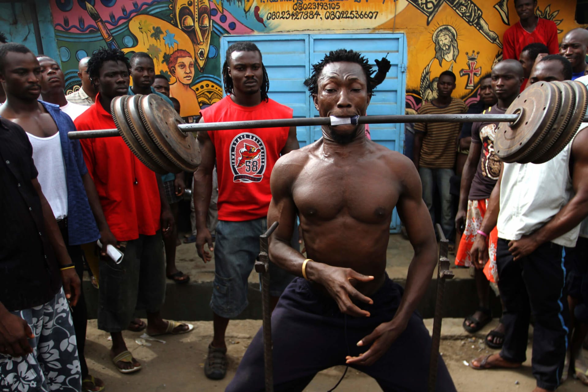 <p>People watch as a man lifts a bar containing 50 kg (110 lbs) iron weights with his mouth at an open gym in Lagos on March 27, 2010. </p>
