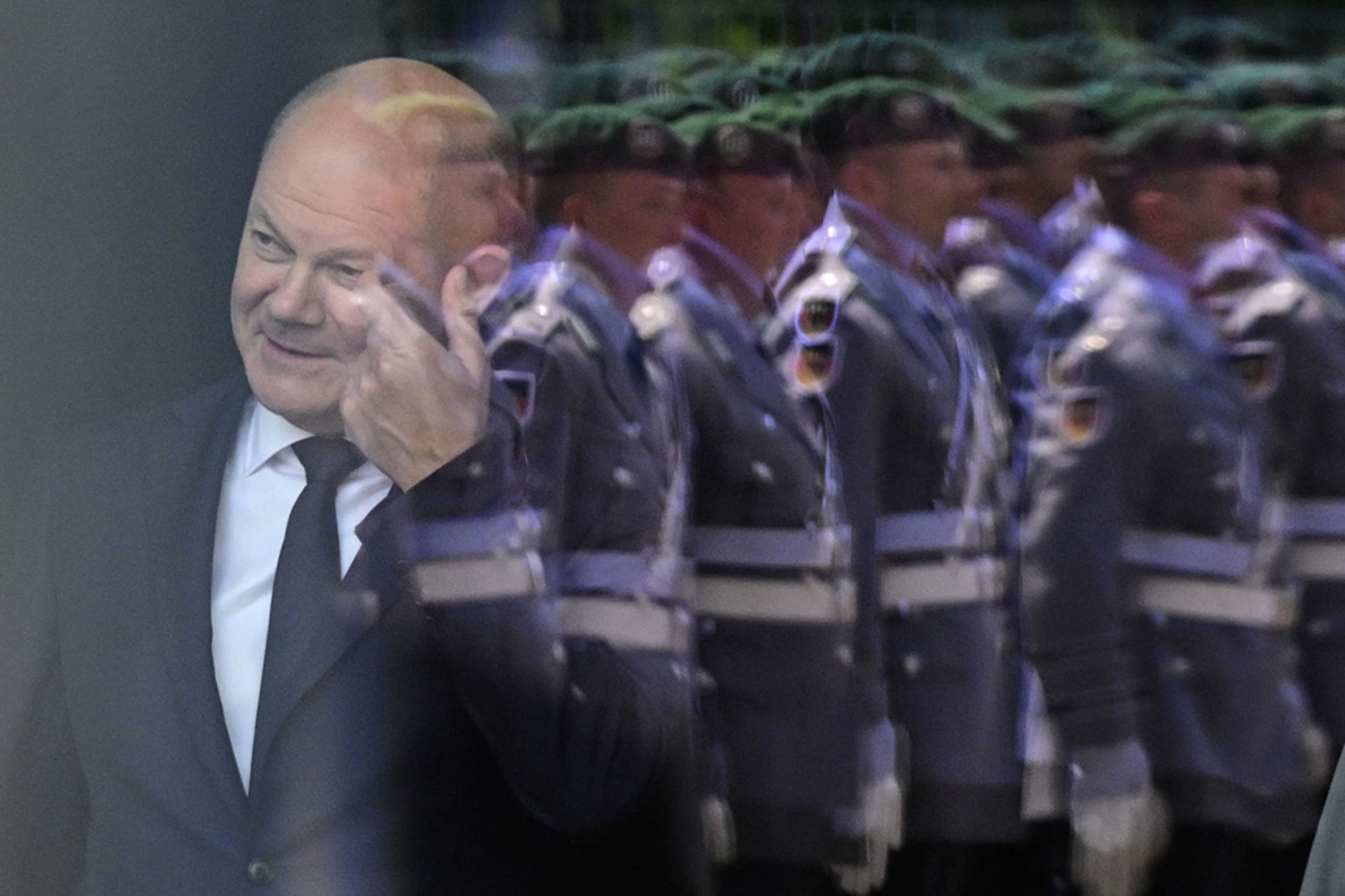 <p>German Chancellor Olaf Scholz stands behind a window in Berlin featuring a reflection of a military honor guard.</p>
