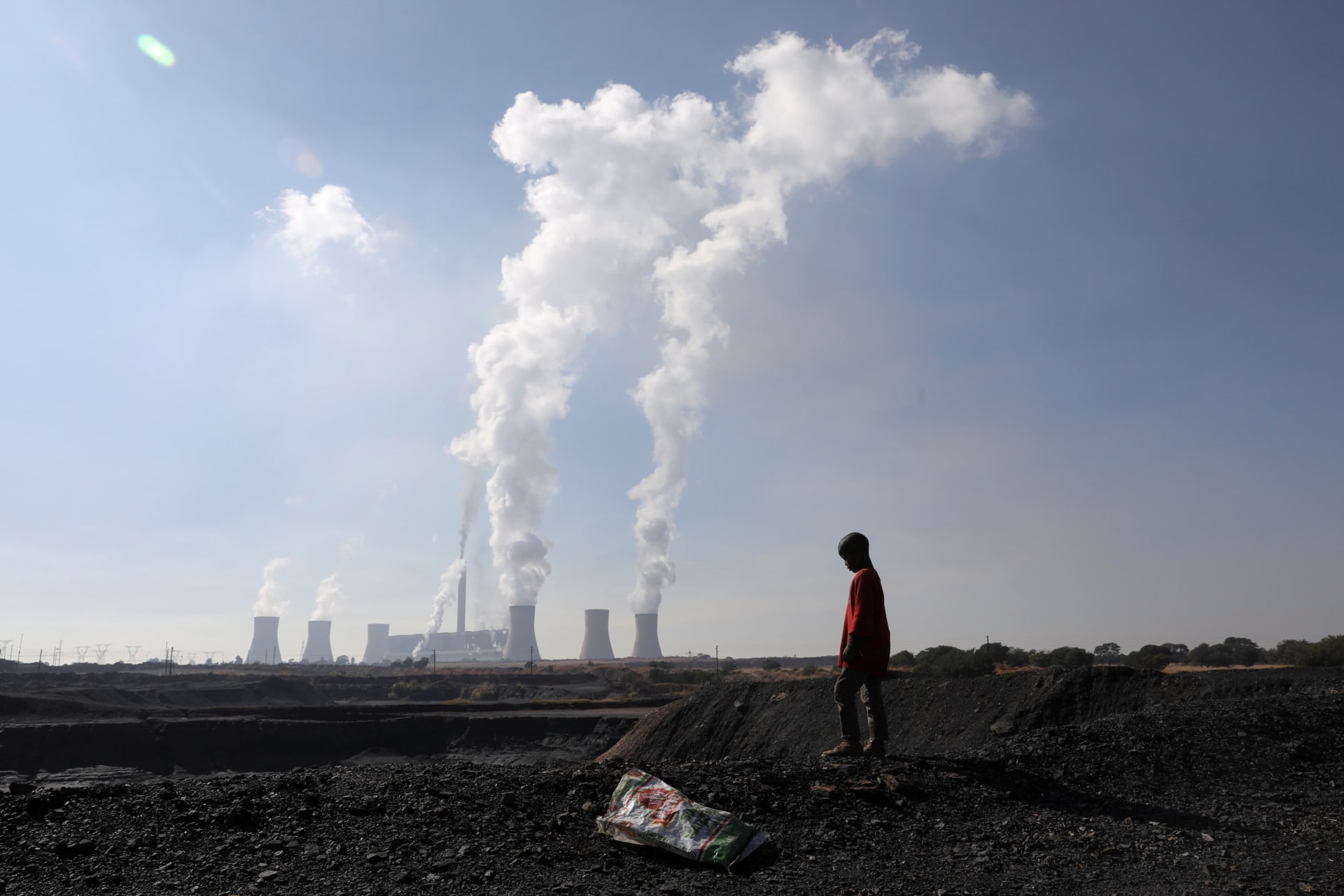 <p>A child collecting chunks of coal looks on at a colliery while smoke rises from the Duvha coal-based power station owned by state power utility Eskom, in Emalahleni, Mpumalanga province, South Africa on June 2, 2021.</p>

