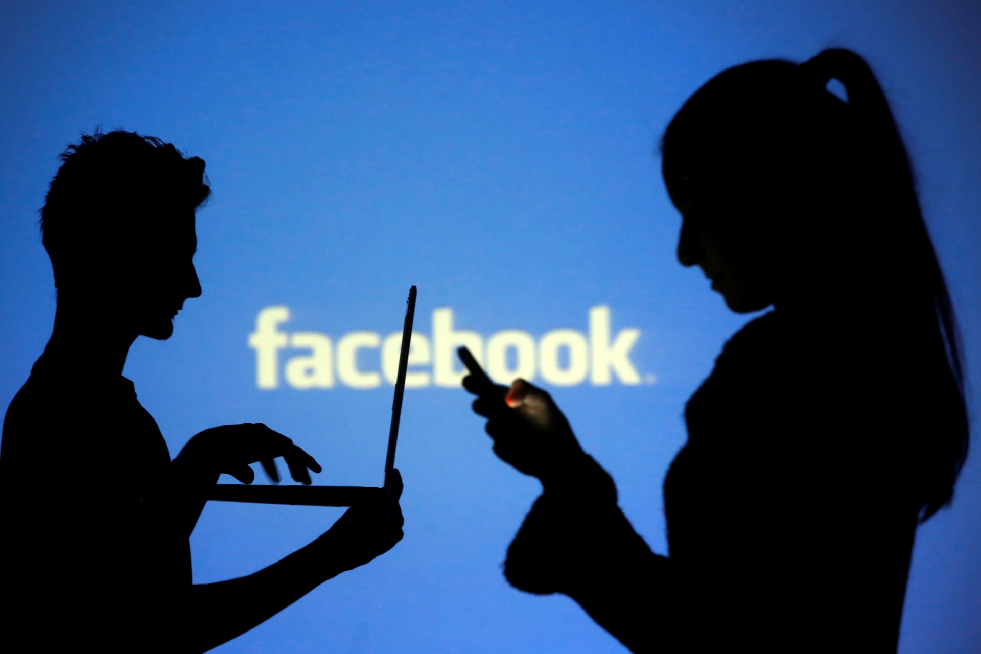 <p>People are silhouetted as they pose with laptops in front of a screen projected with a Facebook logo.</p>
