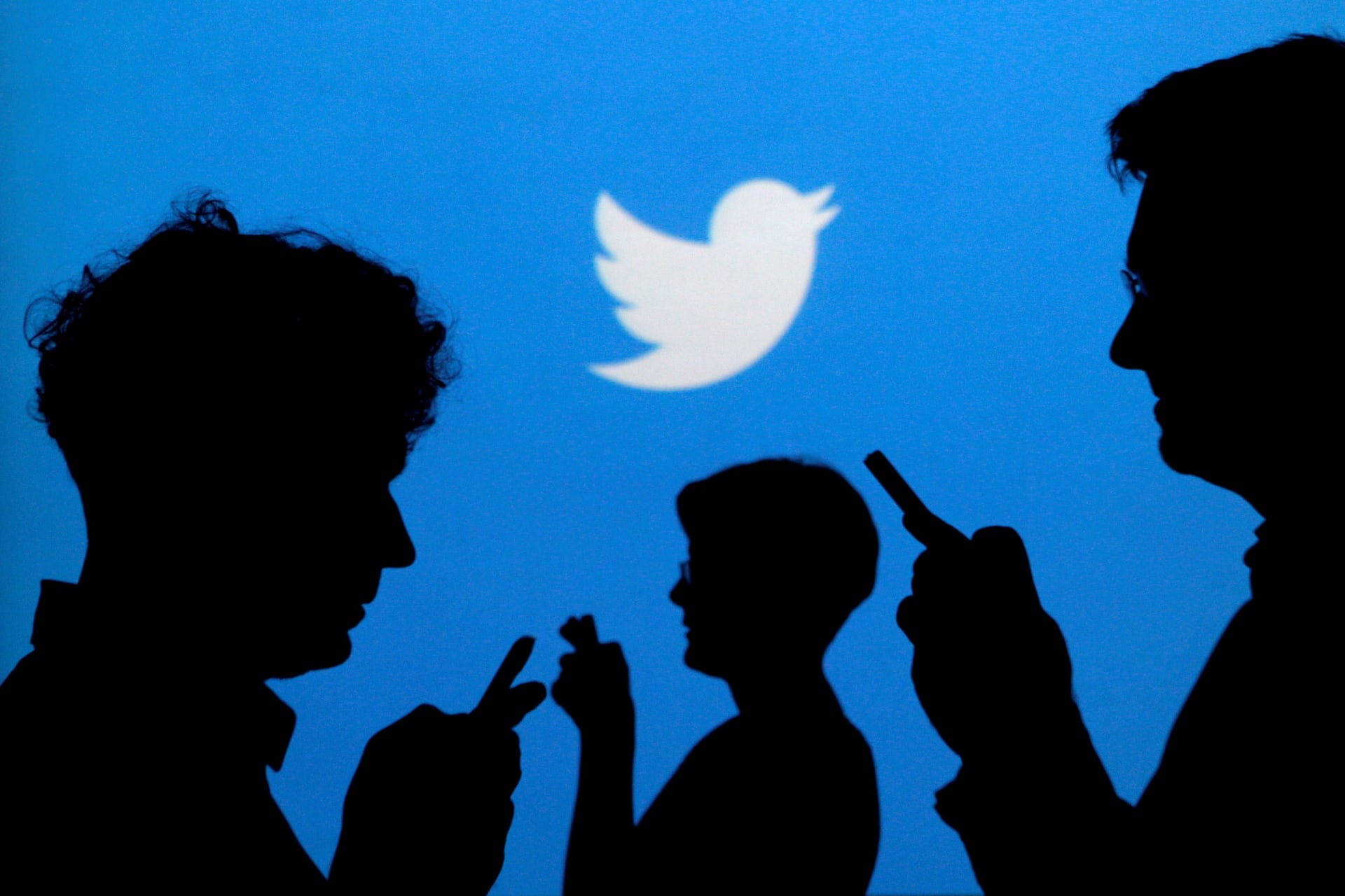 <p>People holding mobile phones are silhouetted against a backdrop projected with the Twitter logo.</p>