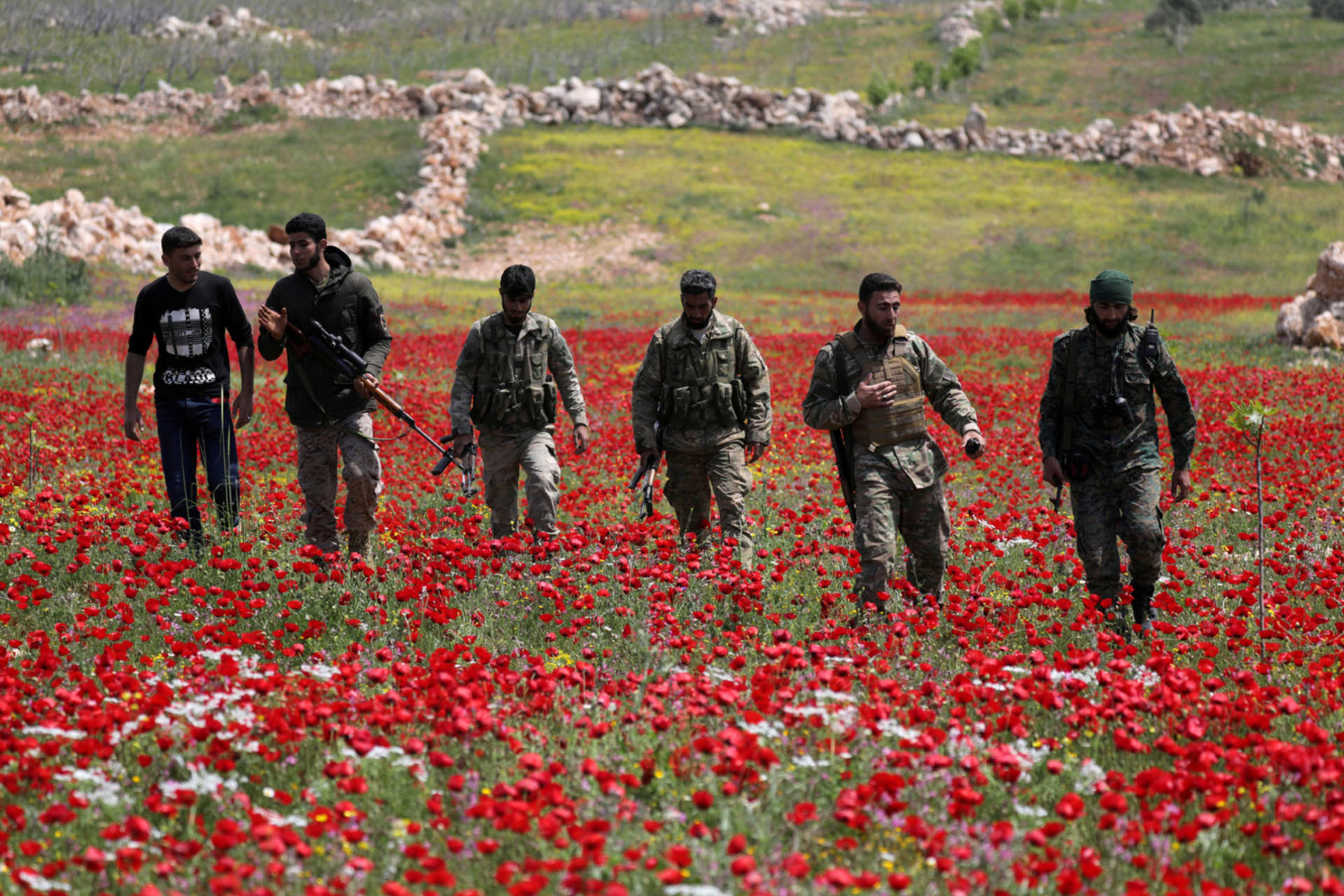 <p>Turkey-backed Syrian rebel fighters walk in a field of flowers in Idlib, Syria, on April 15, 2020.</p>
