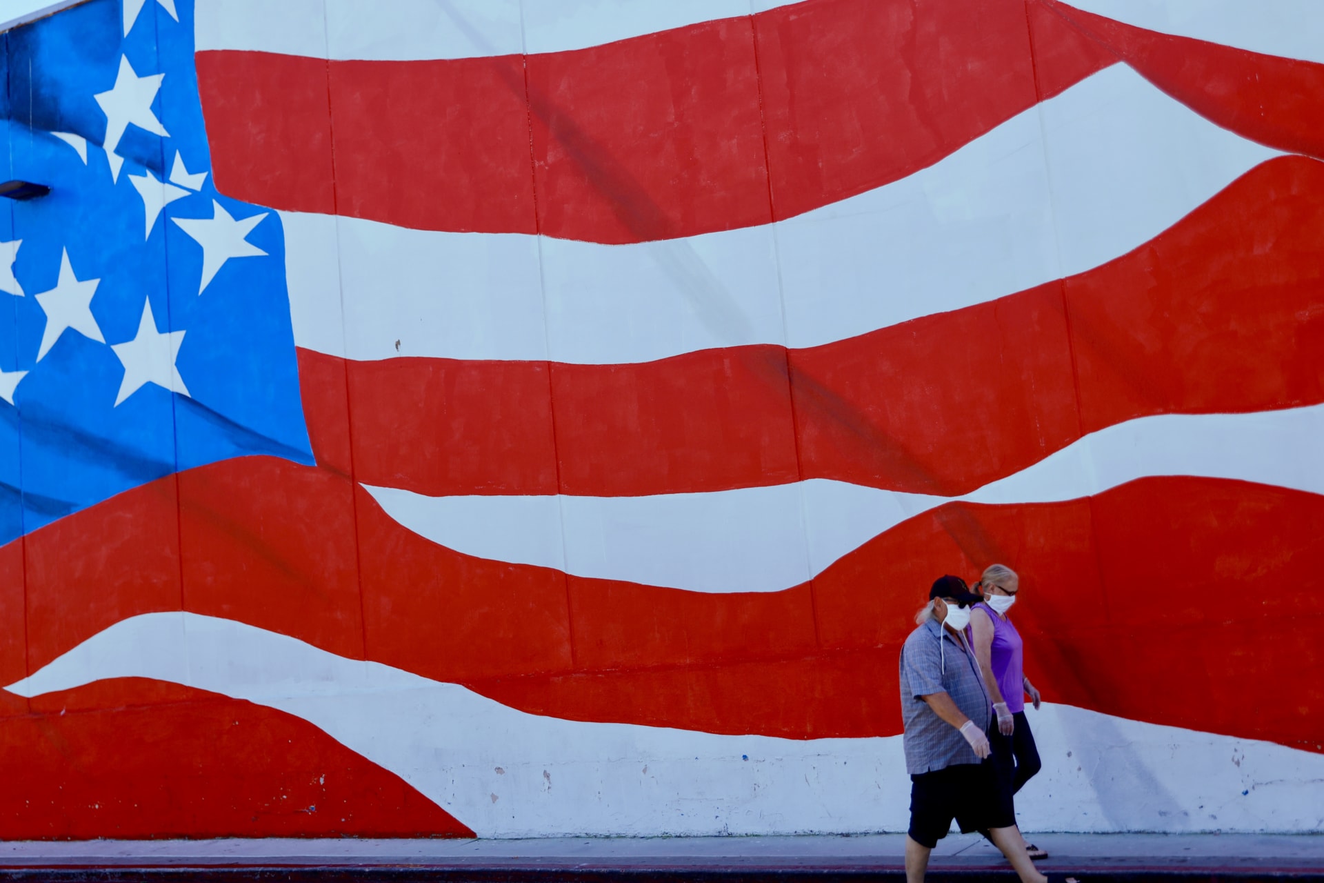 <p>Two people in masks walk by a mural of the U.S. flag in Ocean Beach, California.</p>
