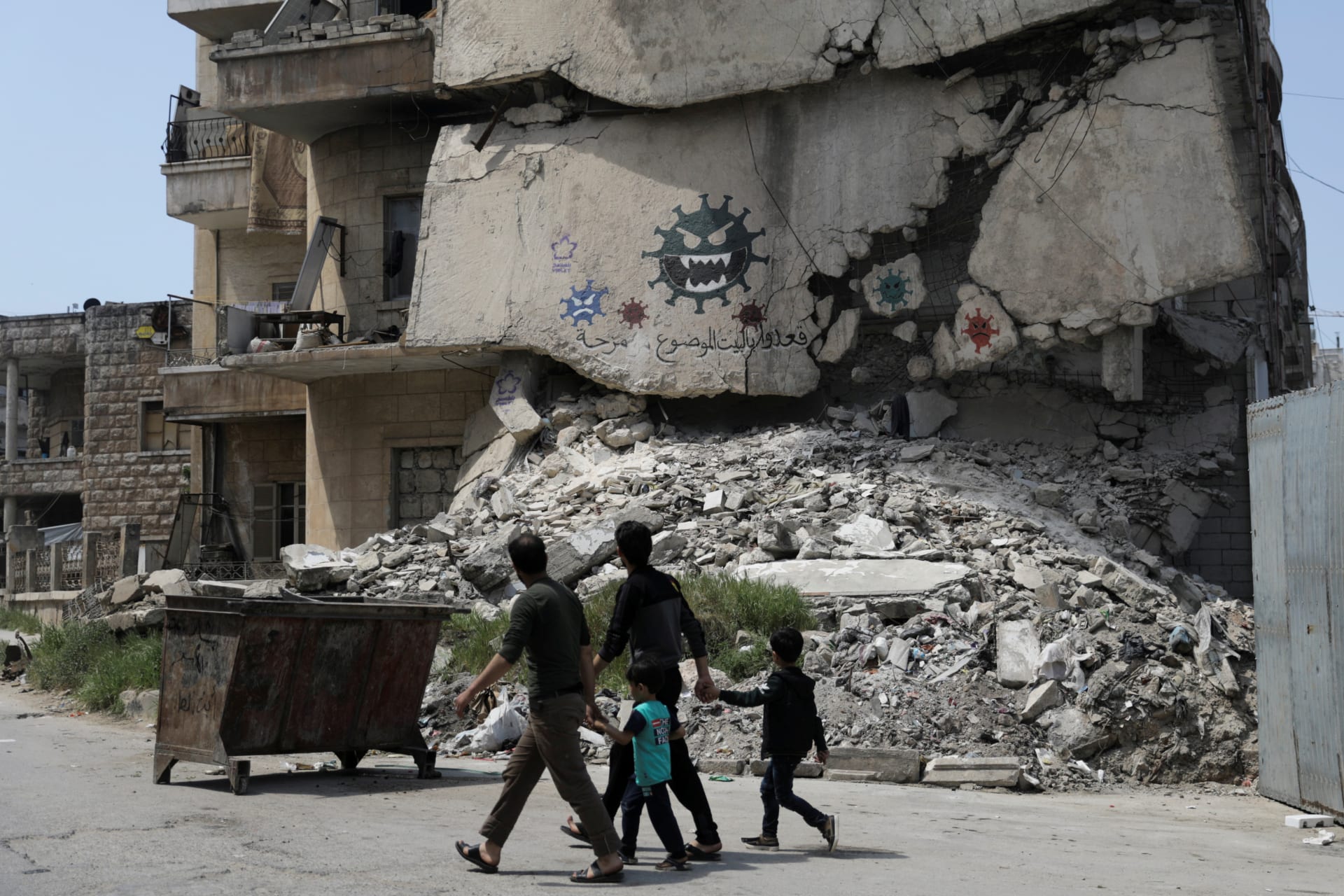 <p>People walk by a damaged building on which illustrations of the novel coronavirus have been spray painted in Idlib, Syria, on April 18, 2020.</p>
