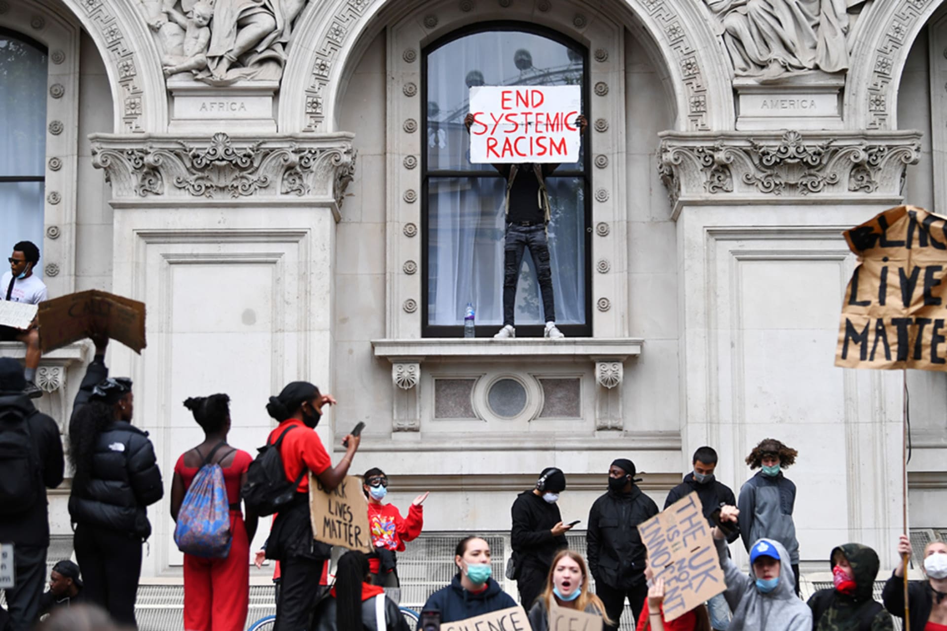 <p>A man holds a banner over the window ledge of a building during a Black Lives Matter protest in London following the death of George Floyd.</p>

