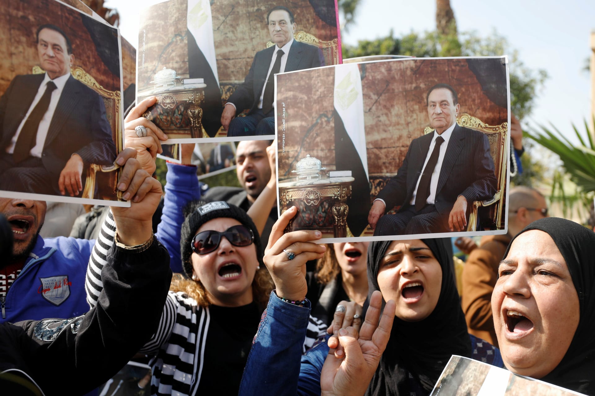 <p>Supporters of former Egyptian President Hosni Mubarak hold his photos near the main gate of a cemetery during his burial ceremony, east of Cairo, Egypt February 26, 2020.</p>
