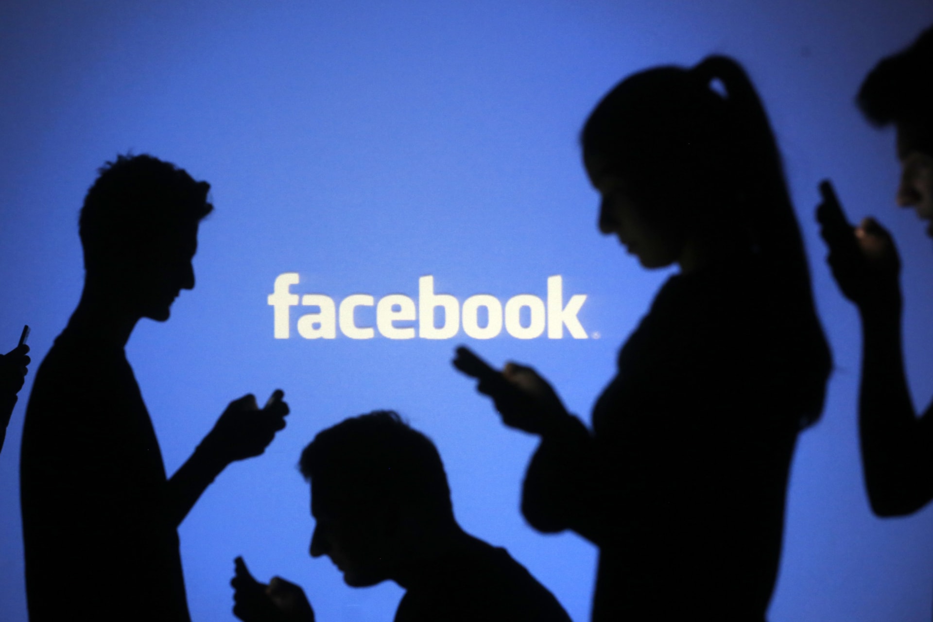 <p>People are silhouetted as they pose with mobile devices in front of a screen projected with a Facebook logo</p>

