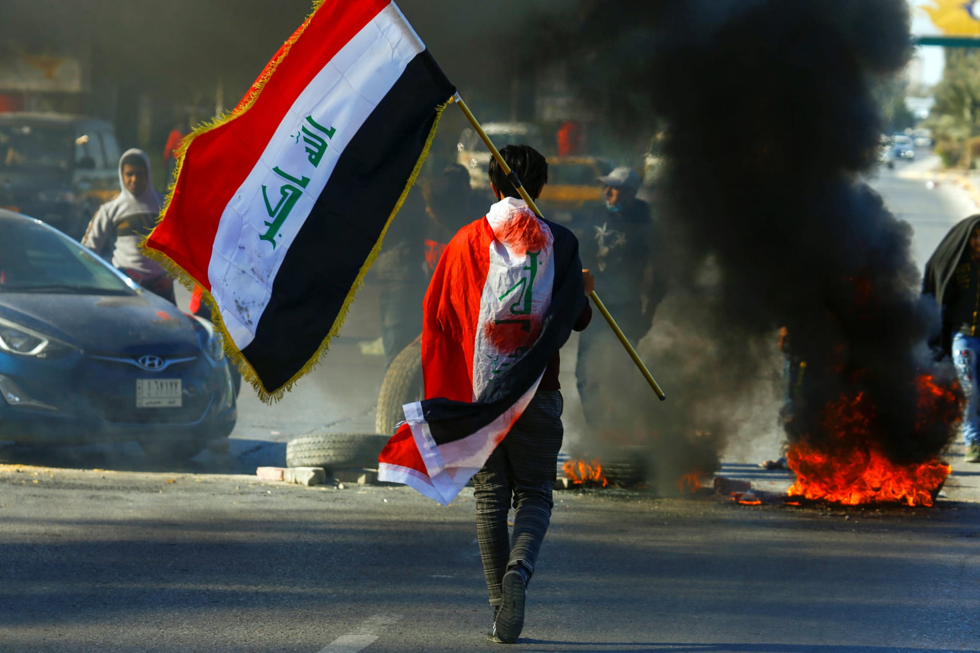 <p>A demonstrator carries an Iraqi flag as he walks near burning tires, during ongoing anti-government protests in Najaf, Iraq January 12, 2020.</p>

