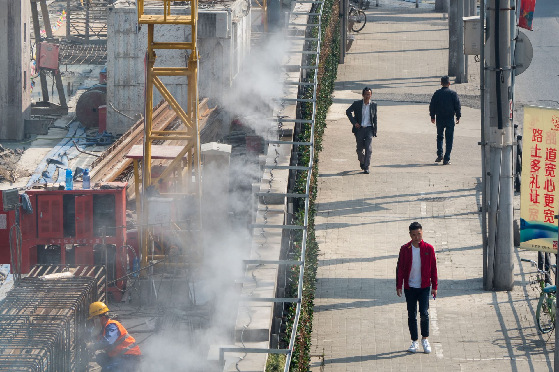 <p>Men walk by a construction site, separated by a wall installed with water misting system for dust control, in Shanghai, China April 8, 2019.</p>

